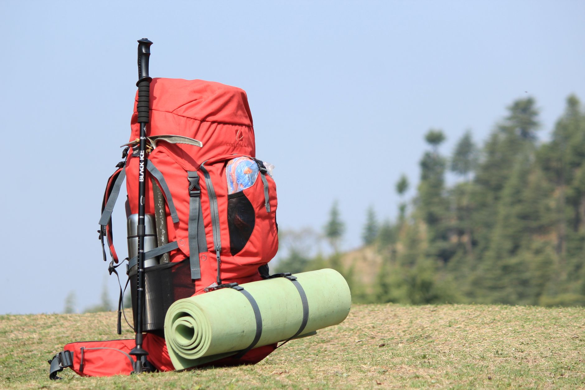 Sac à dos rouge, bâton de marche et tapis de sol posés dans l'herbe au bord d'un sentier