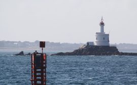 Petit phare blanc à lanterne rouge sur un îlot rocheux, avec au premier plan une balise métallique rouge surmontée d'un voyant carré
