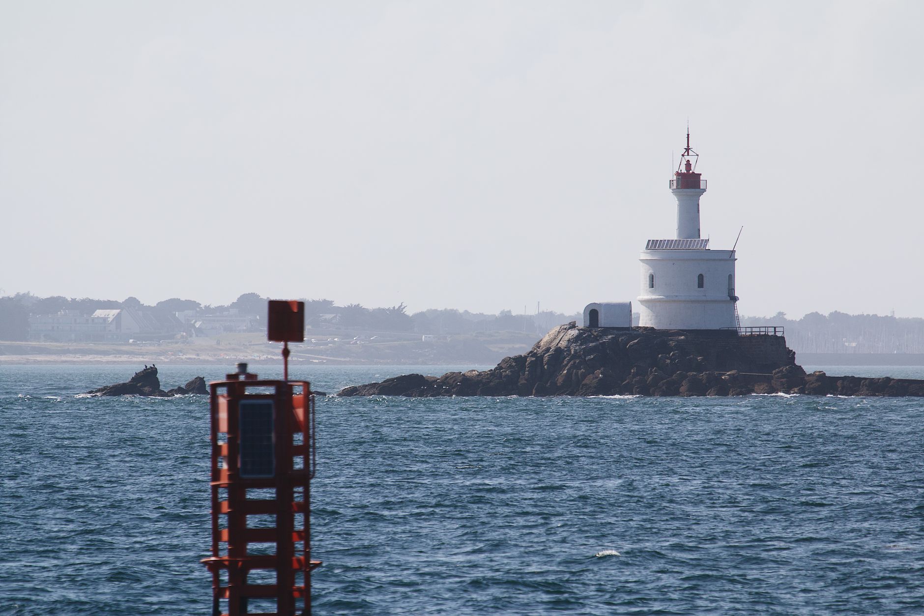 Petit phare blanc à lanterne rouge sur un îlot rocheux, avec au premier plan une balise métallique rouge surmontée d'un voyant carré