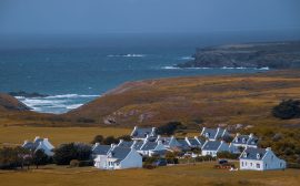 Hameau de maisons blanches aux toits d'ardoise au pied d'une colline couverte de lande, face à une côte rocheuse et à la mer