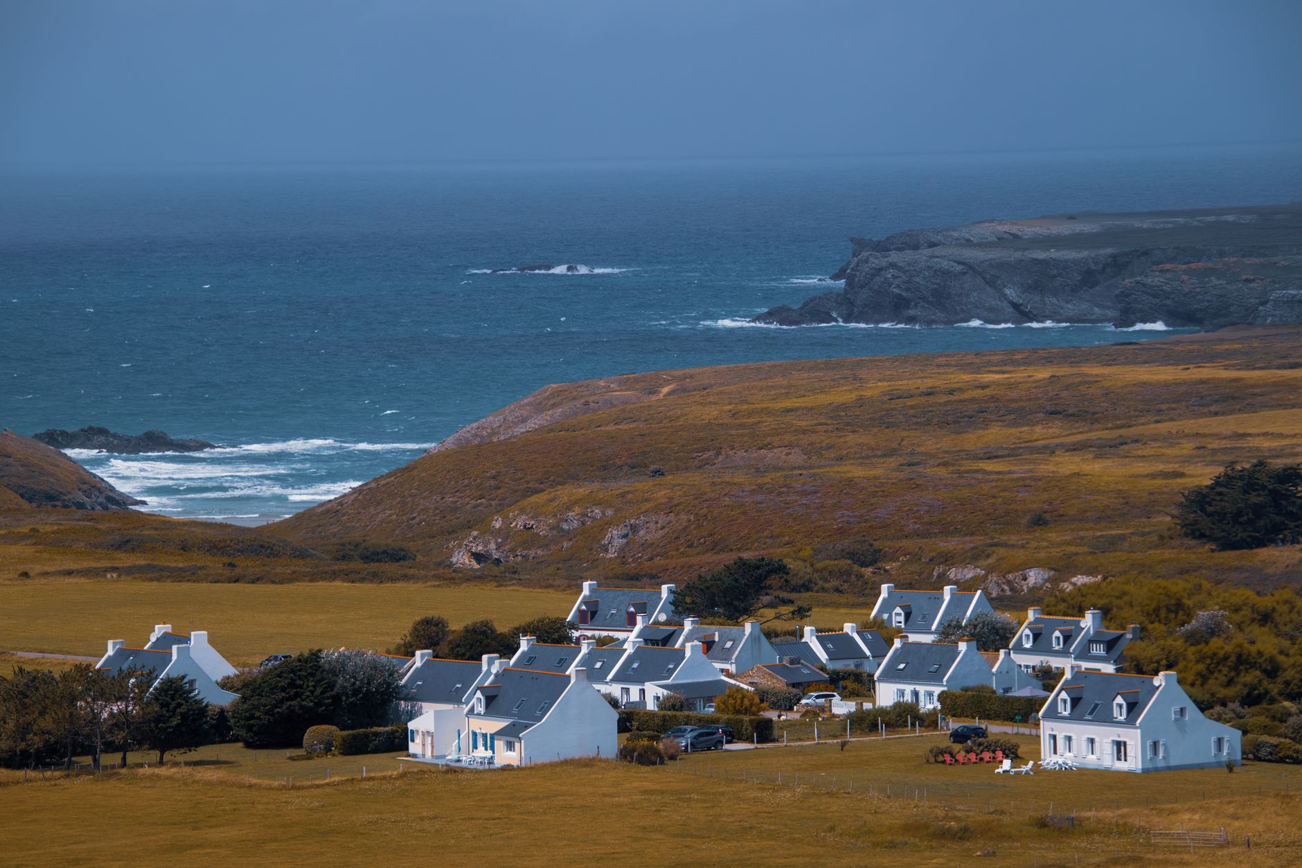 Hameau de maisons blanches aux toits d'ardoise au pied d'une colline couverte de lande, face à une côte rocheuse et à la mer
