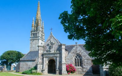 Église bretonne en granite avec haute flèche ajourée, pignon sculpté et porche, entourée de pelouse sous un ciel bleu