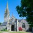Église bretonne en granite avec haute flèche ajourée, pignon sculpté et porche, entourée de pelouse sous un ciel bleu