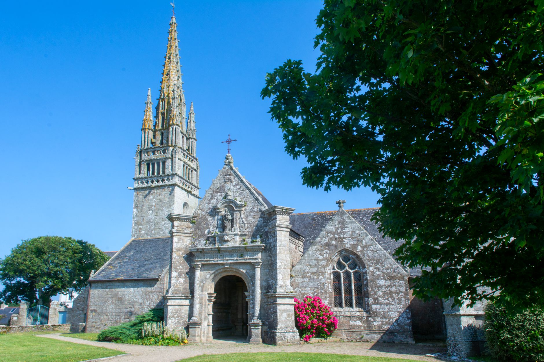 Église bretonne en granite avec haute flèche ajourée, pignon sculpté et porche, entourée de pelouse sous un ciel bleu