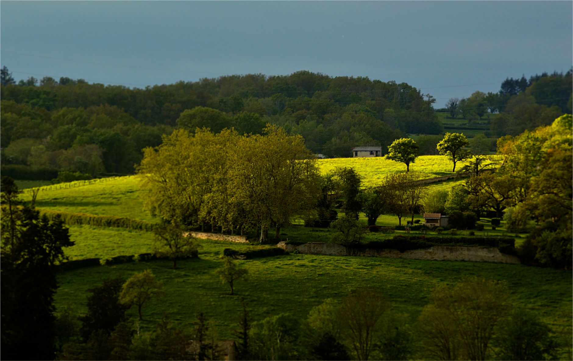 Campagne vallonnée au printemps : prairies séparées par des haies et des rangées d'arbres, murets de pierre et petites maisons, sous un ciel d'orage