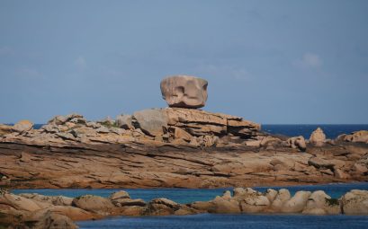 Gros bloc de roche rosée arrondi, posé en équilibre sur une dalle rocheuse, au-dessus d'une mer bleue et d'autres rochers arrondis