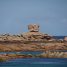 Gros bloc de roche rosée arrondi, posé en équilibre sur une dalle rocheuse, au-dessus d'une mer bleue et d'autres rochers arrondis