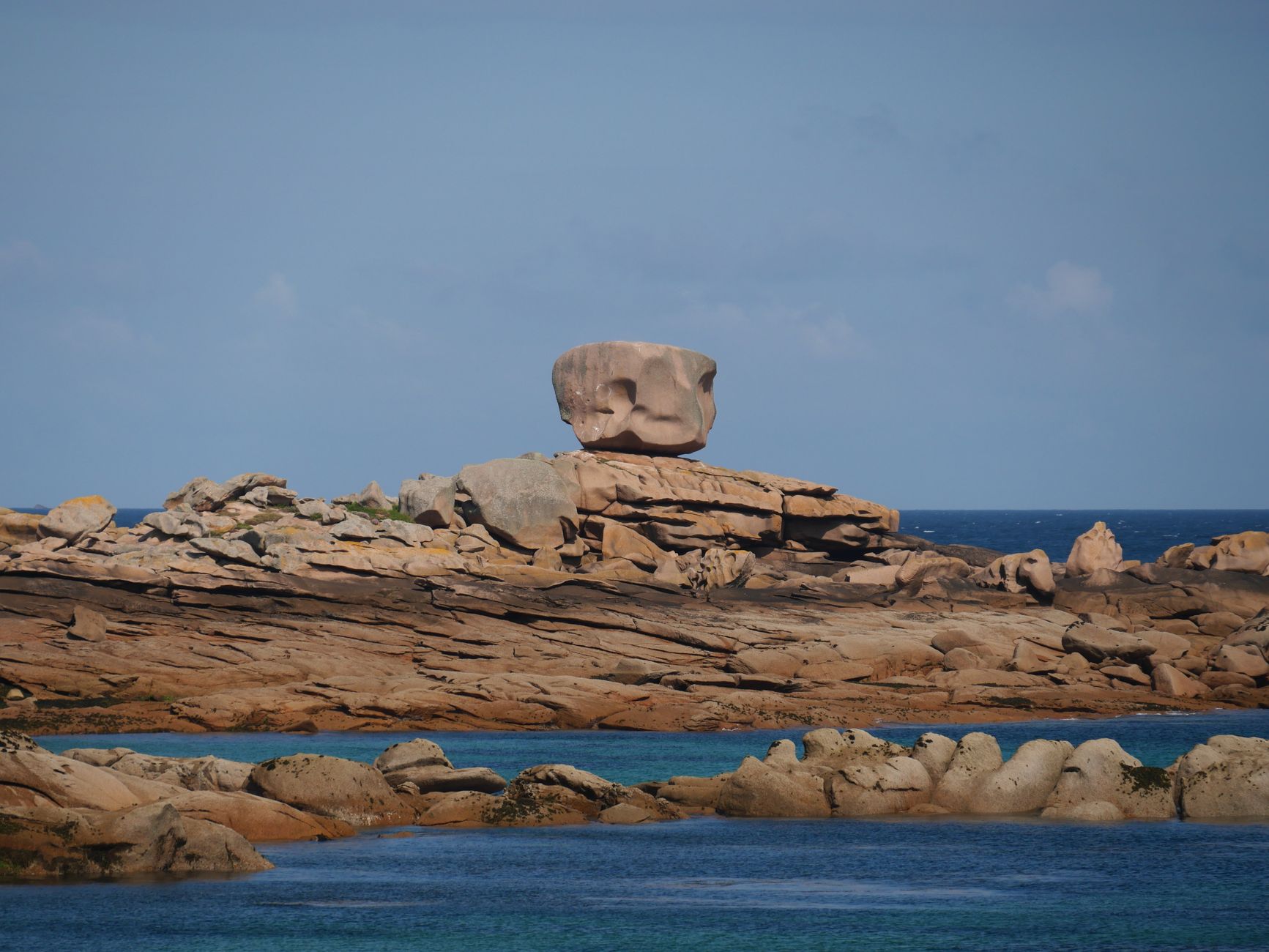 Gros bloc de roche rosée arrondi, posé en équilibre sur une dalle rocheuse, au-dessus d'une mer bleue et d'autres rochers arrondis