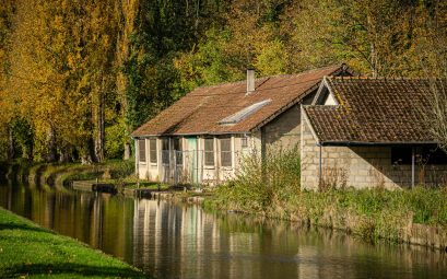 Canal bordé d'arbres aux feuillages d'automne, avec une rive herbeuse et une ancienne maison en pierre au bord de l'eau