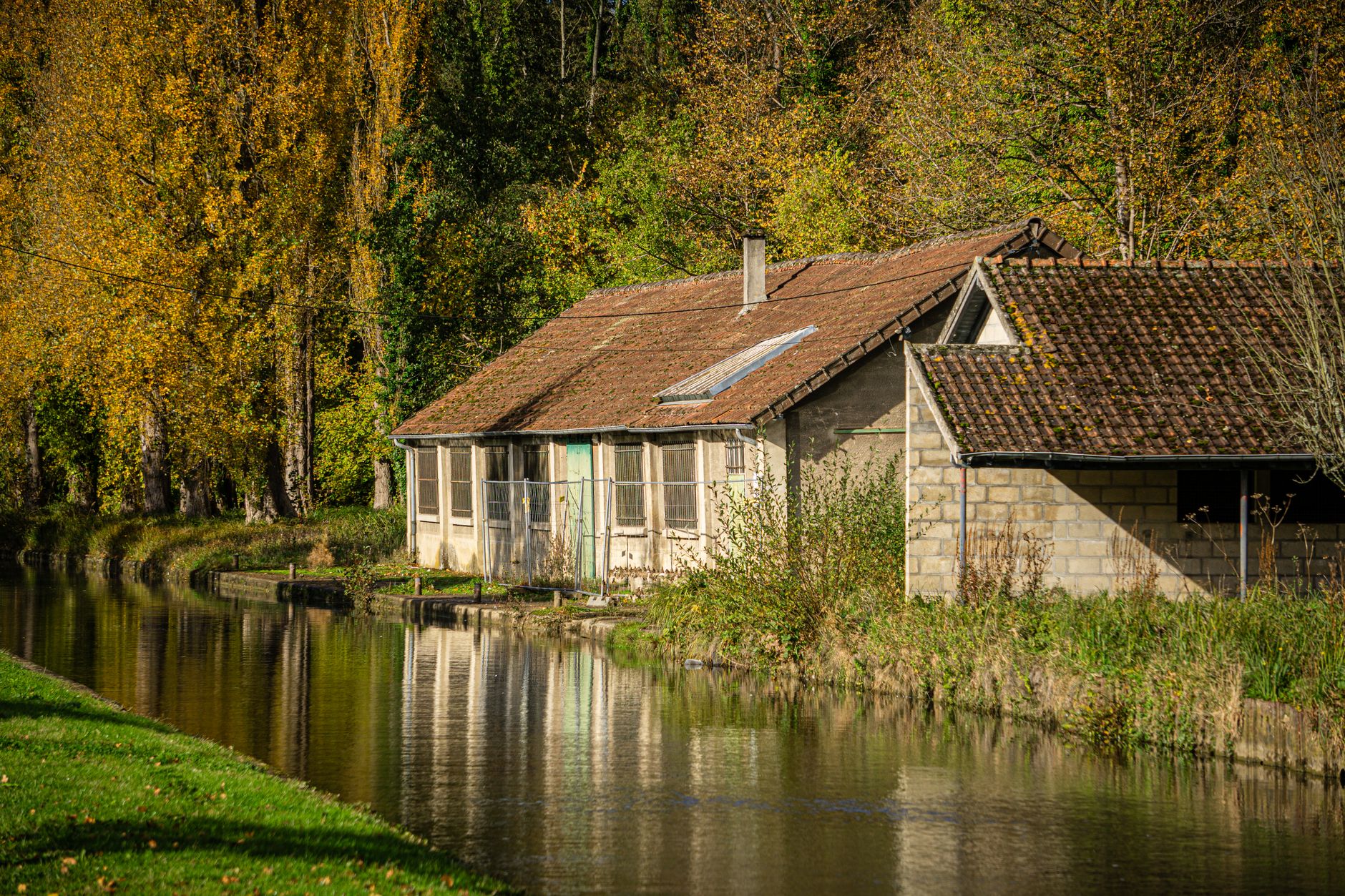 Canal bordé d'arbres aux feuillages d'automne, avec une rive herbeuse et une ancienne maison en pierre au bord de l'eau