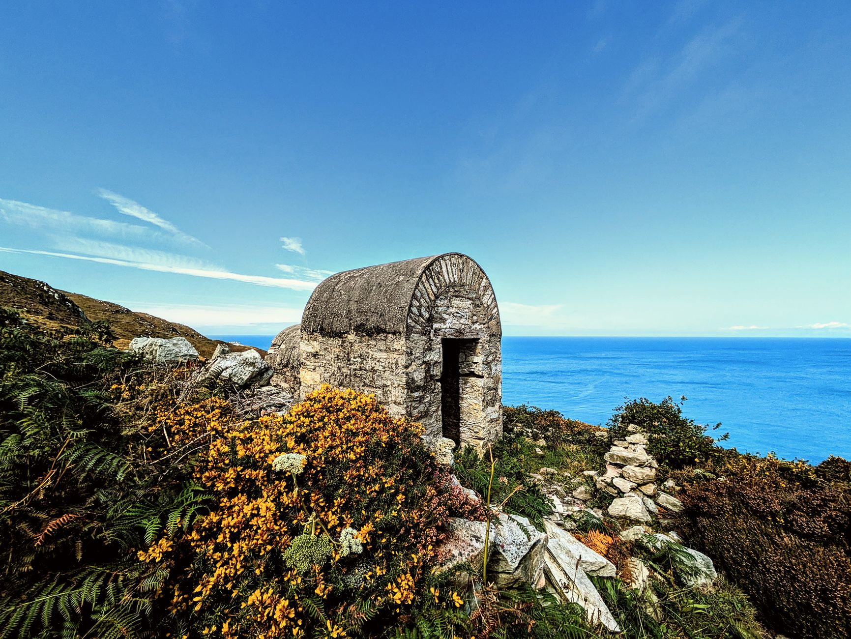 Petit abri de pierre voûté au sommet d'une côte, entouré d'ajoncs en fleurs et de fougères, face à la mer